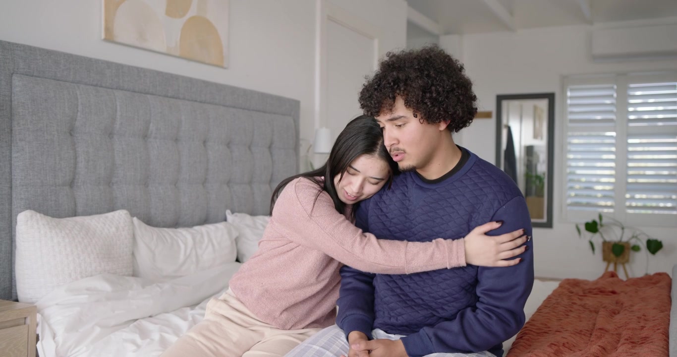 Couple sitting on bed in bedroom, woman speaking then reaching, hugging to comfort worried man