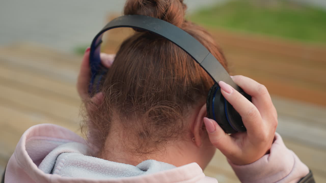 Close up back view of girl in pink hoodie gently lifting headphones to head with relaxed motion and blurry background showing wooden surface and lush greener