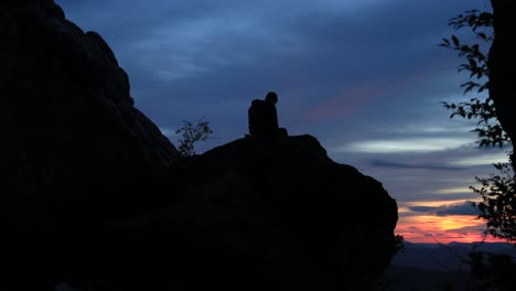 A-man-sits-in-silhouette-on-a-large-boulder-at-the-Dragon's-Tooth-rock-monolith-on-the-Appalachian-Trail-in-Virginia-and-watches-the-sunrise