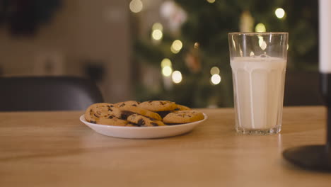 Camera-Focuses-On-An-Glass-Of-Milk-And-A-Plate-Full-Of-Cookies-On-An-Empty-Table-In-A-Room-Decorated-With-A-Christmas-Tree