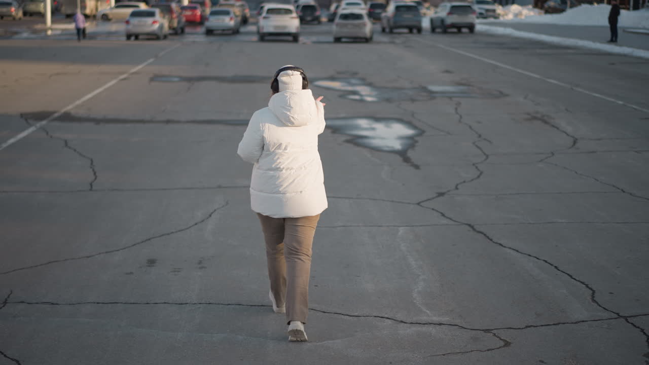 Back view of cheerful young woman in white jacket and headphones swaying rhythmically across cracked urban pavement with scattered water patches, joyfully expressing herself on cold winter day
