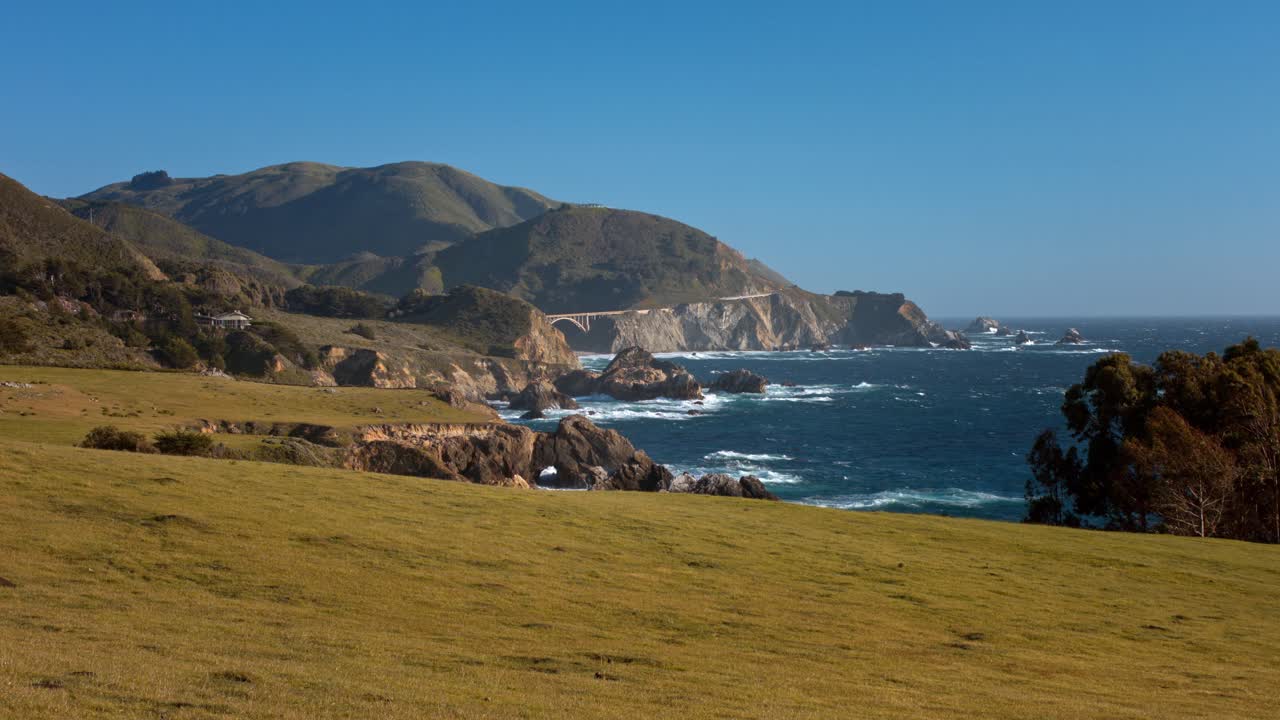 Coastal hills meet ocean with rocky shoreline under clear blue sky. Vibrant green fields lead to the water, creating a peaceful, natural landscape in sunny weather