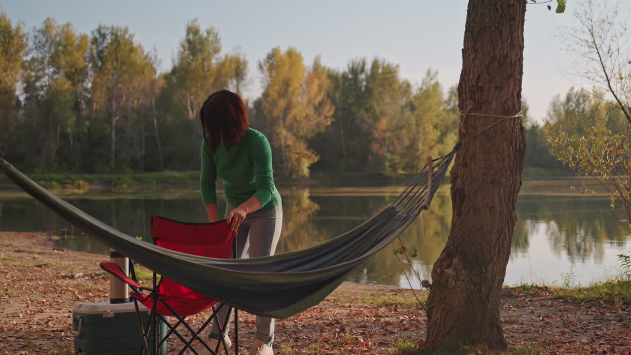 Kvinde sætter campingstol ved søen, stille scene ved søbredden med hængekøje bundet mellem træer, rød klapstol, køleboks ved bredden, efterårslys reflekteres i vandet, fredfyldt soloafslapning og naturskøn