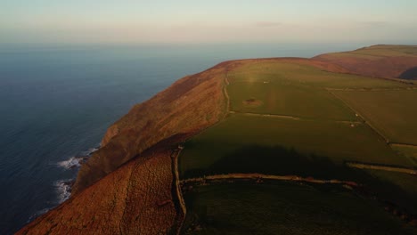 Aerial View of Sunset Lit Farmed Fields in Rural Devon with Coastline Sea Waves Crashing Below as Camera Pans Around Revealing Bristol Channel Ocean. Drone Coastal Nature Footage 4K.