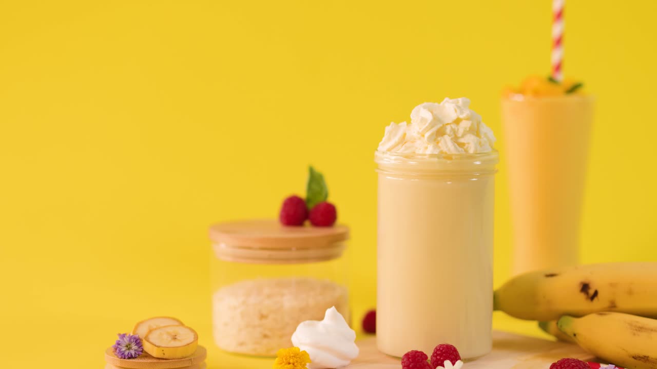 Primo piano statico di un frullato di banana su sfondo giallo, una mano aggiunge fette di banana e panna montata