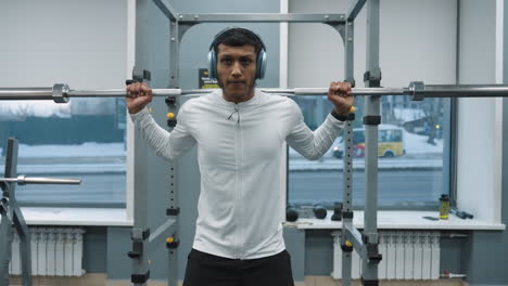 young athletic man carrying heavy barbell on shoulder while performing squats inside gym with natural light coming through window and blurry view of moving car outside during strength training
