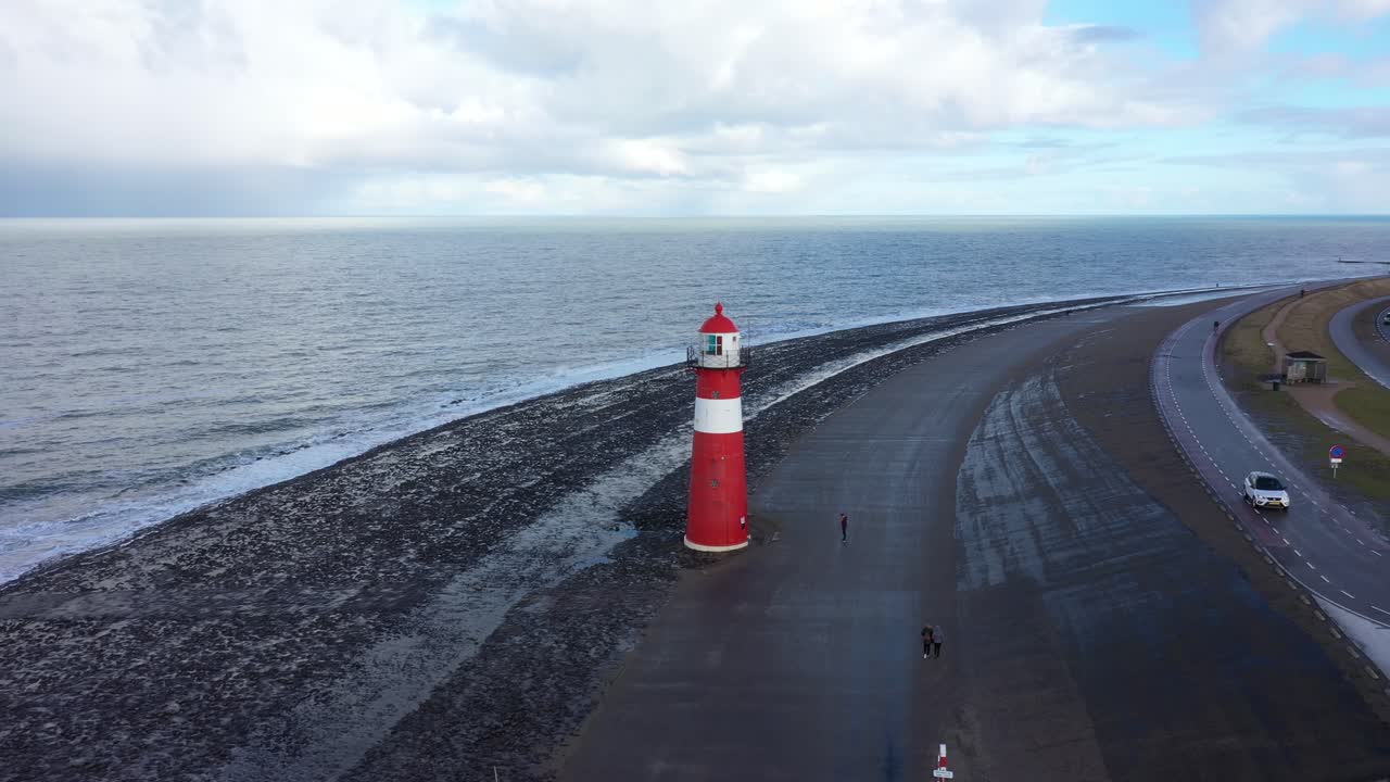 Drone shot of a lighthouse by the Dutch coastline, with a guy standing next to it, flying to the left and panning right.