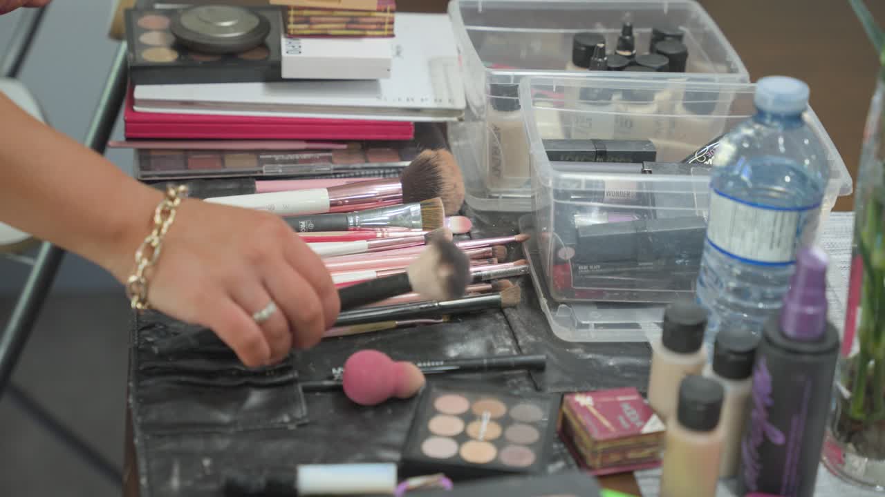 A high-angle shot of a professional makeup station featuring an array of brushes, eyeshadow palettes, foundations, and organizers as a stylist selects a tool for application