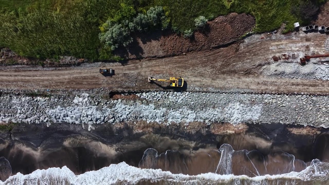Lavori di costruzione su un argine di fiume con macchinari pesanti che movimentano terra e rocce in una zona rurale durante il giorno.