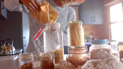 Feliz-Mujer-Afroamericana-Vertiendo-Pasta-En-Un-Frasco-En-Una-Cocina-Soleada,-En-Cámara-Lenta