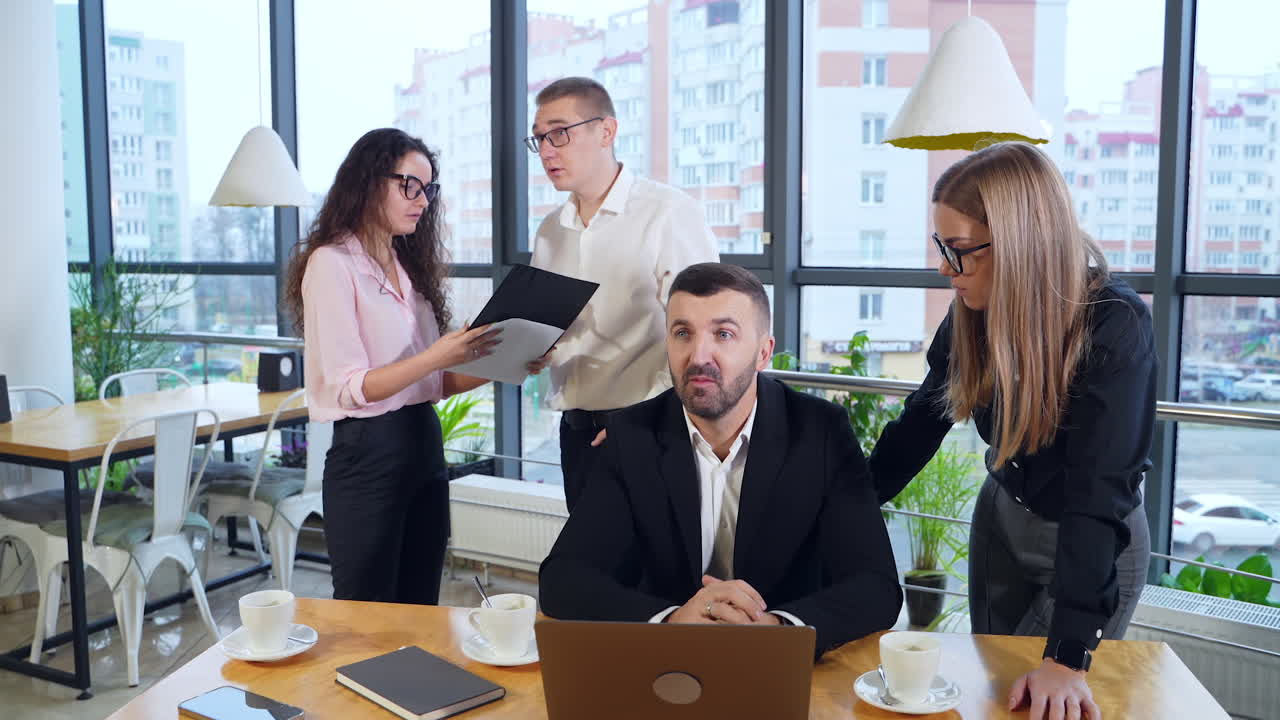Employer sits at the desk in front of laptop and woman standing beside him and discussing issues on computer. Two colleagues speaking about chart in woman's hands.