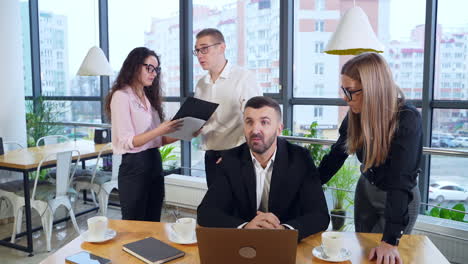 employer sits at the desk in front of laptop and woman standing beside him and discussing issues on computer. two colleagues speaking about chart in woman's hands.