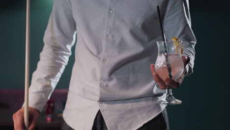 person in white shirt and grey trousers, face not visible, holding cue stick and glass of lemon drink with black straw. ice cubes and lemon slice float in glass, creating a refreshing look