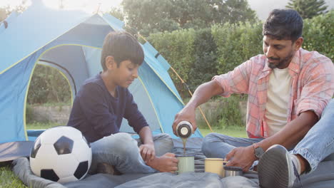 camping with son, indian father pouring drink from thermos near tent and soccer ball
