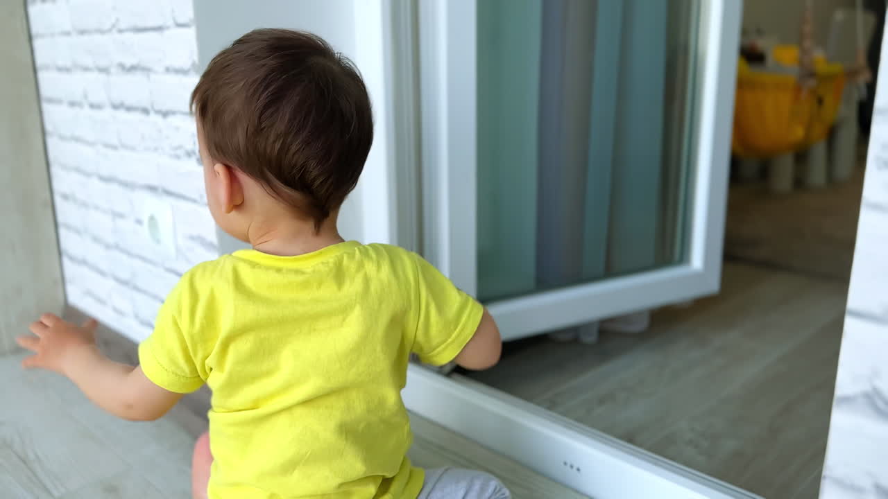 Sweet baby boy sits on the balcony touching electric socket on the brick wall. Kid moves to the room but then returns back.