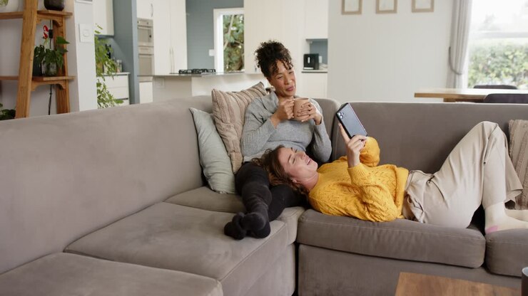 Diverse female friends tapping tablet to show content, sipping from mug and smiling on couch