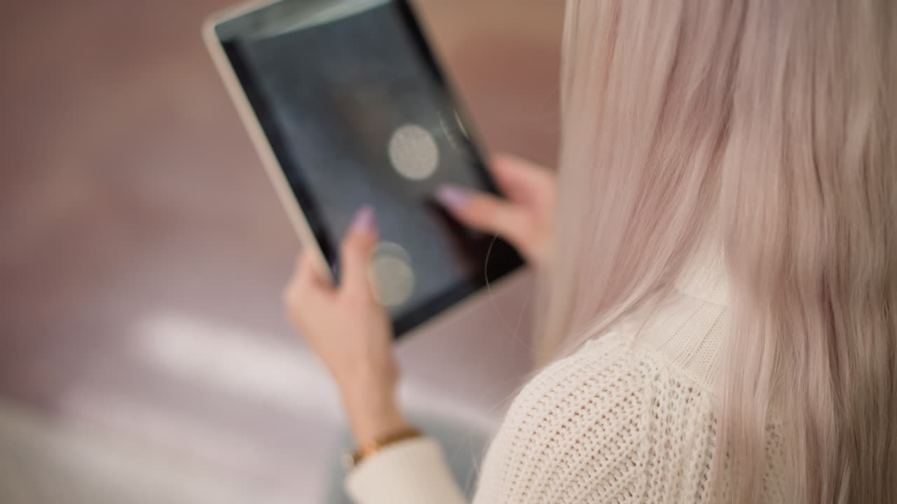 overhead view of elegant young lady with long flowing hair seated on bench in bright mall hall tapping tablet screen with manicured nails focusing on digital browsing, blurred bokeh background lights