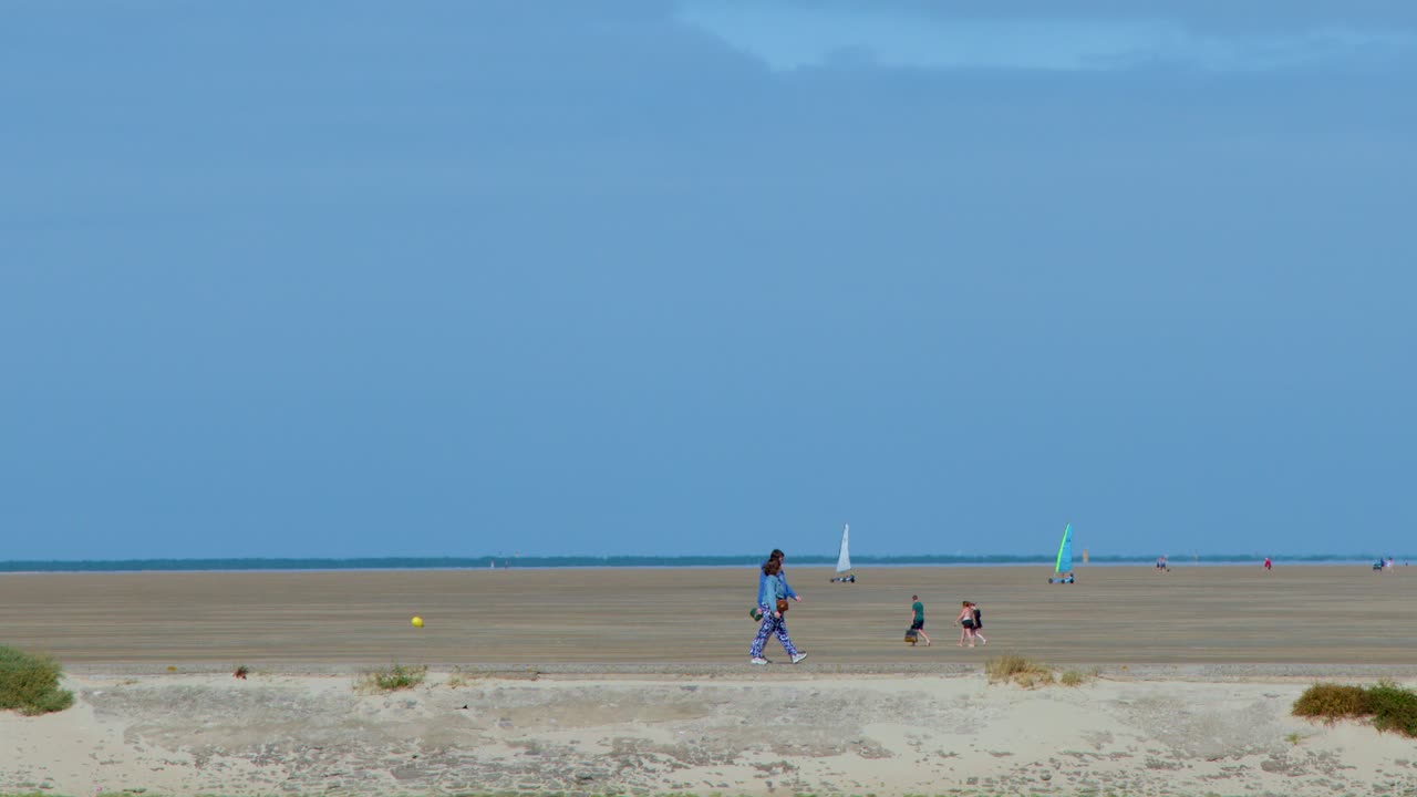 Tourists stroll on a wide sandy beach under clear daylight, distant sailboats and calm atmosphere