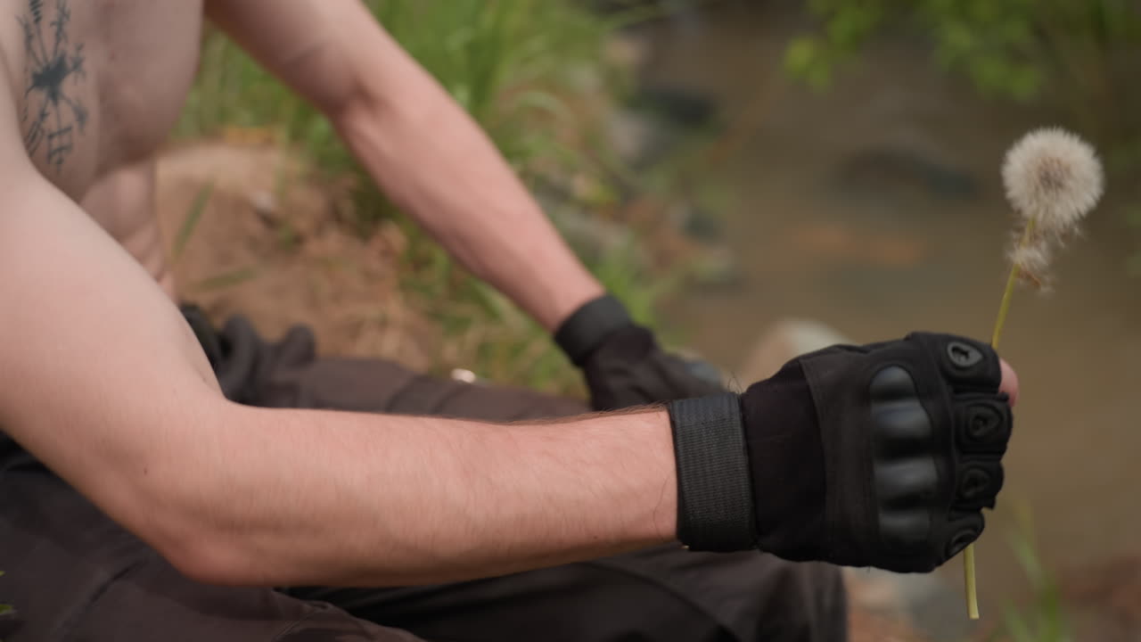Close-up of shirtless warrior wearing fingerless glove and headband, bending over on grassy terrain while supporting elbow with knee, near muddy water and scattered stones, suggesting outdoor focus