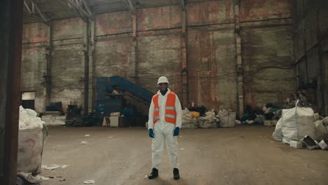 retrato de un hombre feliz y confiado con color de piel negra en ropa de protección blanca y un chaleco naranja que se para y cruza los brazos en su pecho en una enorme planta de reciclaje y clasificación de residuos