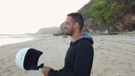 slow motion of young beautiful white man on the beach smiling and wearing helmet in hand searching for new adventure and beautiful beach for surfing and tropical destination with ocean in background