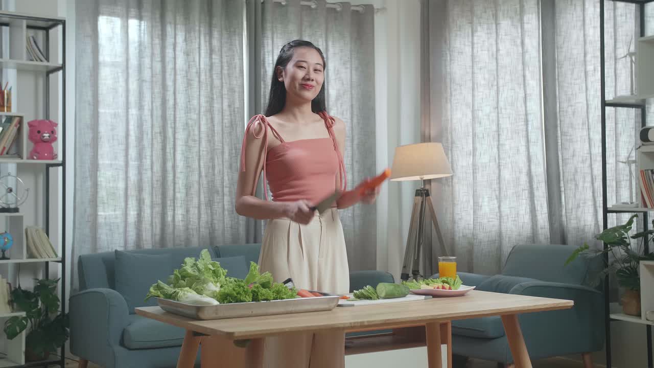 집에서 카메라에 포즈를 취하기 전에 칼을 들고 당근을 잘라내는 미소 짓는 아시아 여성