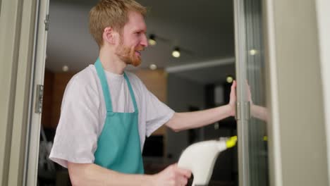 Side-view-of-a-confident-male-blond-cleaner-in-a-white-T-shirt-and-a-gray-blue-apron-washes-the-glass-in-the-door-using-a-vacuum-cleaner-for-windows-in-a-modern-apartment-while-calling-a-cleaning-company