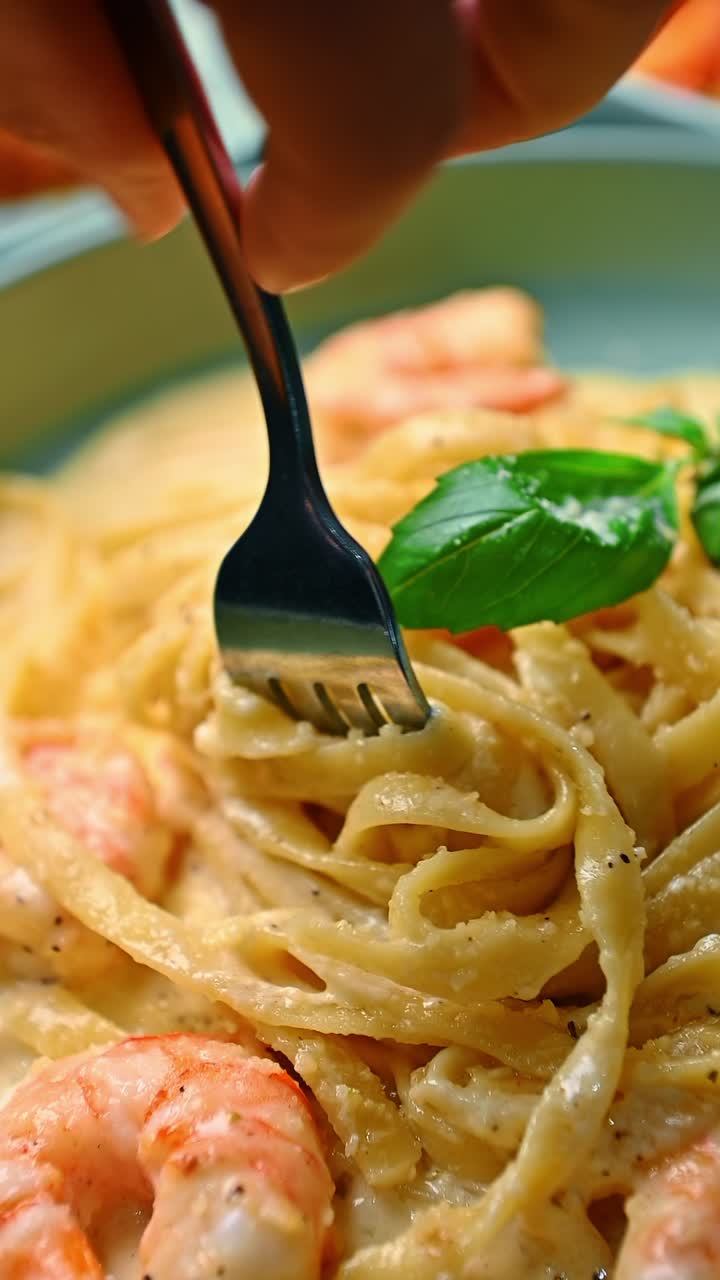 Primo piano di forchetta che avvolge fettuccine con gamberetti. Forchetta d'argento che arrotola lunghe fettuccine in salsa bianca cremosa con gamberetti cotti e basilico verde fresco.