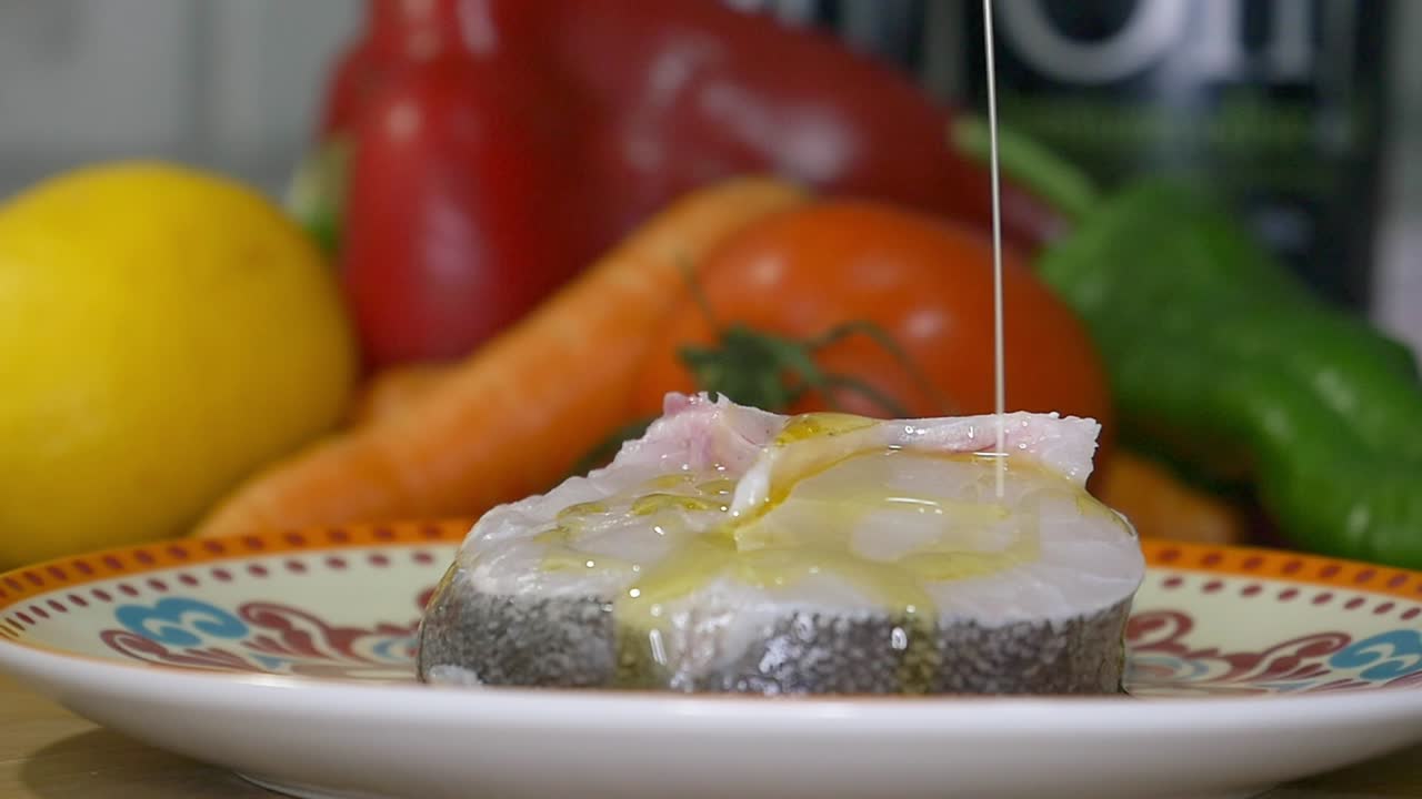 Close-up of a slice of fresh hake on a plate, being drizzled with a fine stream of Extra Virgin Olive Oil (EVOO)