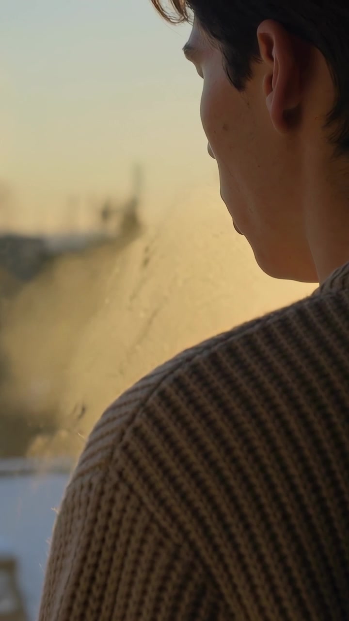 Video verticale: Uomo illuminato dal sole con maglia a coste che si gira guardando oltre un vetro appannato a casa, spazio per il testo.