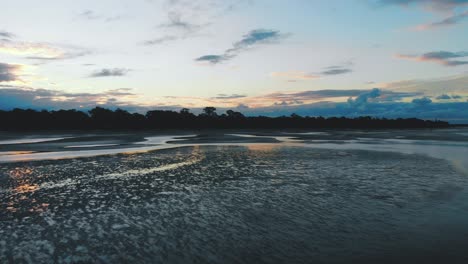 slow push towards the treeline over an amazingly reflective beach from small veins and bodies of water sat from low tide on a warm afternoon whilst the sun spills orange and blue colours into the sky