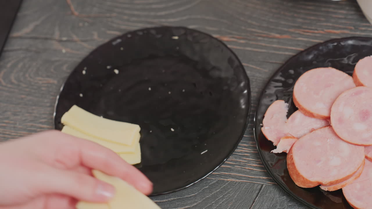 close up of person arranging thin slices of yellow cheese on black ceramic plate next to round slices of sausage on dark wooden surface in kitchen environment with visible hand and food crumbs