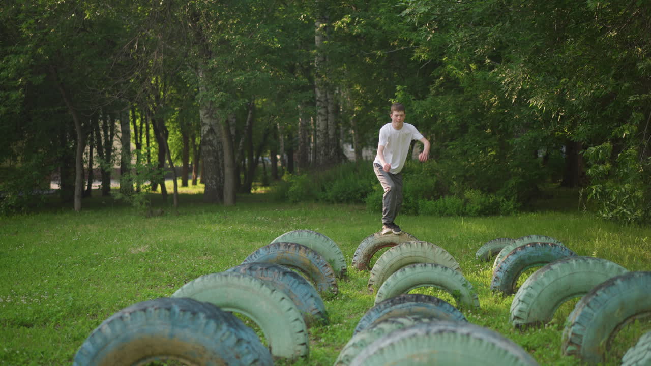 Front view of a young boy in a white shirt and gray pants, jumping on a row of tires in a grassy field, captured in slow motion with green trees in the background