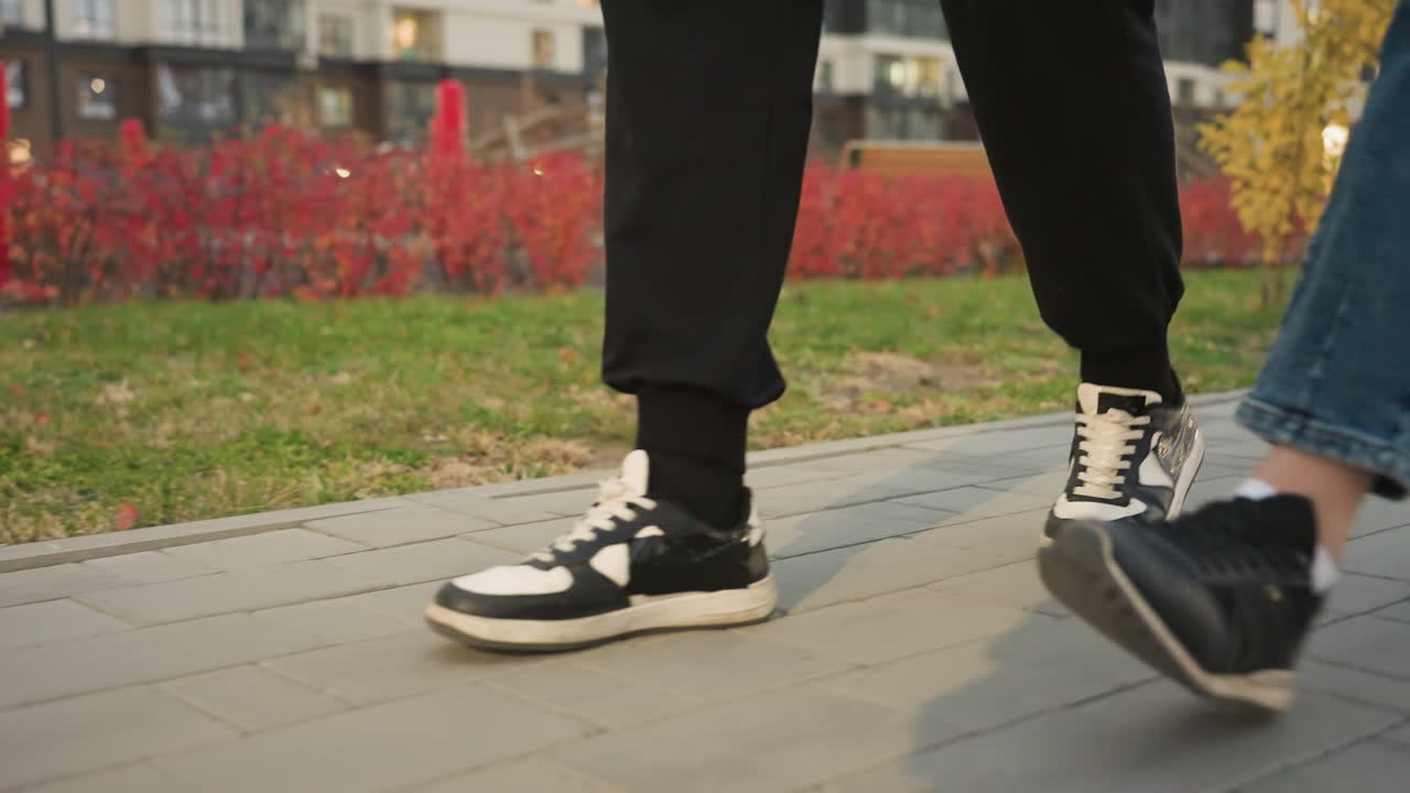 Lower body view of people casually strolling on paved interlocked walkway during evening, wearing sneakers and casual pants, with soft residential building lights glowing in background