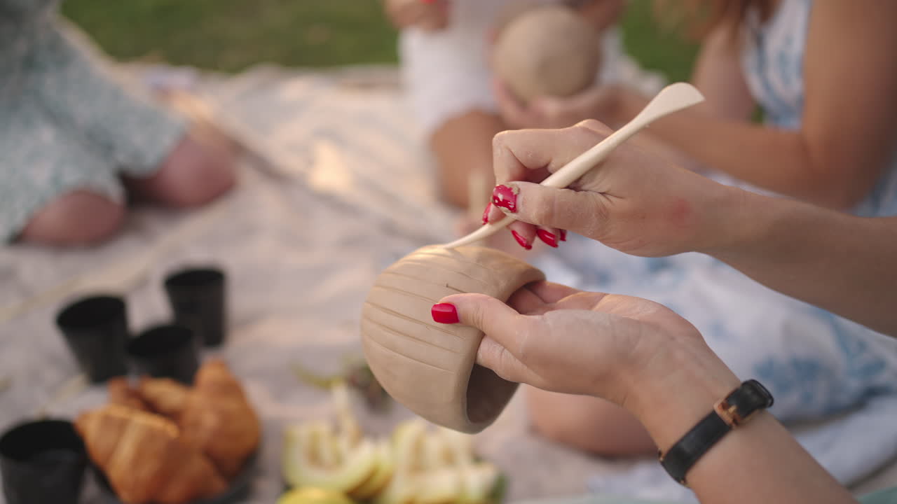 Women concentrate on drawing patterns on clay products with the help of tools in the meadow in nature in the open space. Women's hands decorate the product in close-up.
