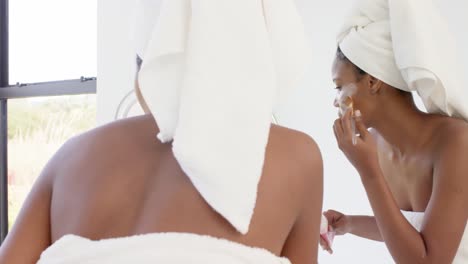 mujer biracial enfocada con toalla en la cabeza haciendo máscara en la cara en el baño, cámara lenta
