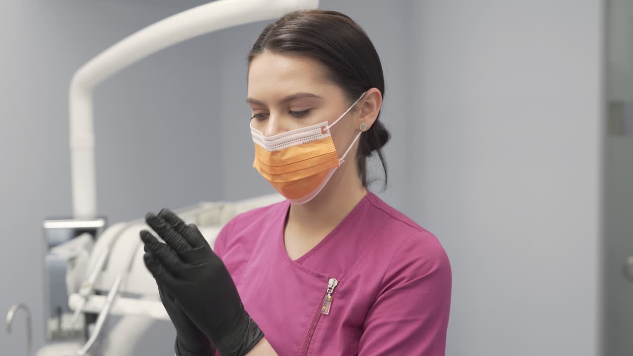 Dentista Feminina em Consultório Odontológico Preparando-se para Exame e Tratamento Usando Luvas Pretas e Corrigindo a Posição da Máscara Enquanto Olha para a Câmera