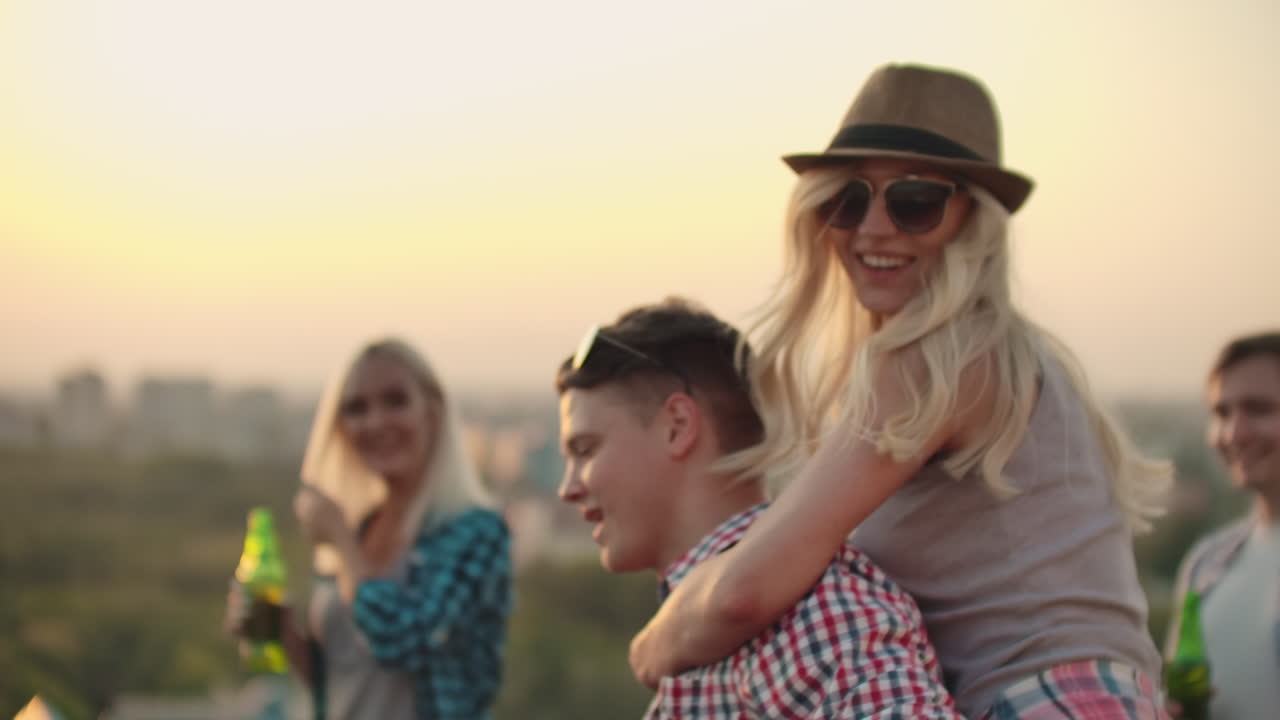 A girl moves in a dance on the back of her boyfriend among their friends. They have fun on the party with friends on the roof.