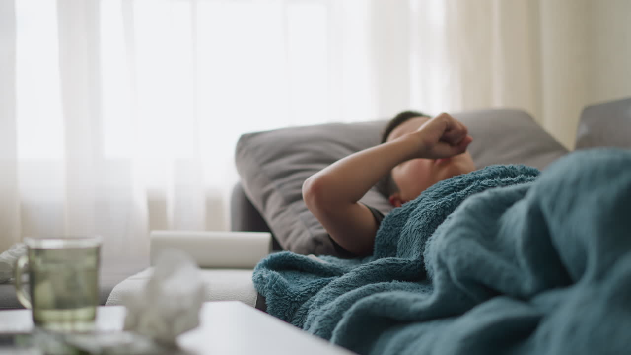 Sick child lying on couch covered with blanket, coughing and resting hand on head, tissue nearby with blurred cup and tissue on table