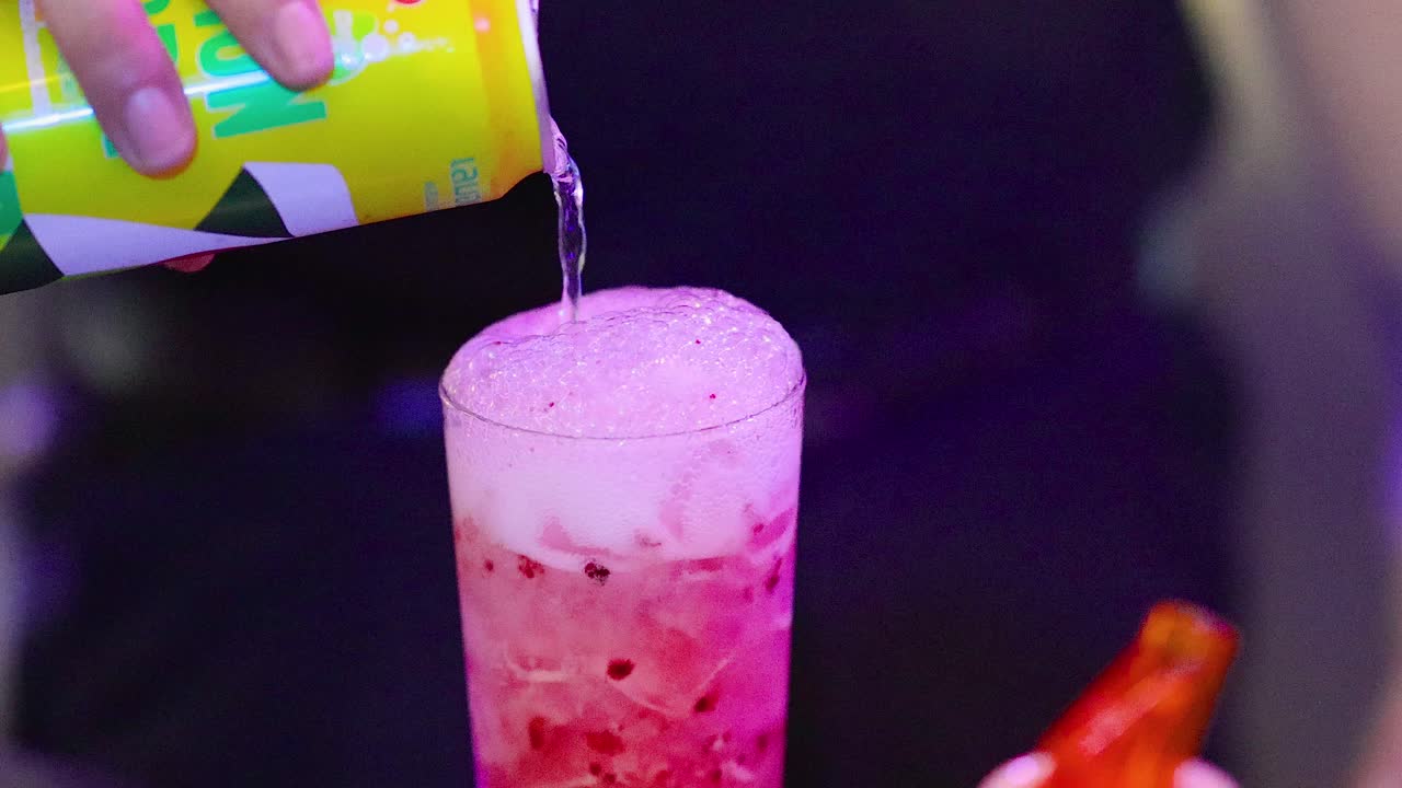 Bartender pours lemon soda into iced cocktail glass under colorful bar lighting, close-up perspective