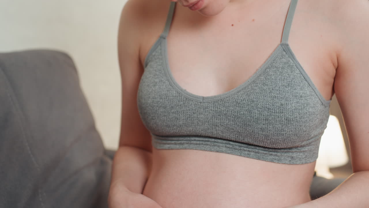 Donna bianca incinta che si tocca la pancia, seduta in un soggiorno illuminato da luce soffusa, indossa un reggiseno sportivo grigio e leggings verdi. Le mani formano un cuore sulla pancia, espressione serena, consapevolezza prenatale e delicata...