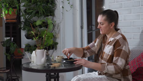 white woman seated on sofa beside glass table opens pot extracts granular substance which she sprinkles onto moist soil in black tray surrounded by potted plants, watering can, gardening tools
