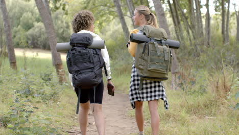 two women with backpacks hike along a forest trail