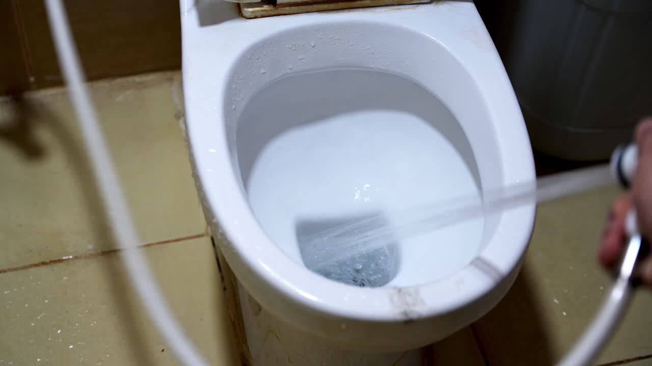 Close-up of water spraying into a white ceramic toilet bowl from a handheld bidet sprayer. Hygienic bathroom cleaning routine using a high-pressure shattaf water jet