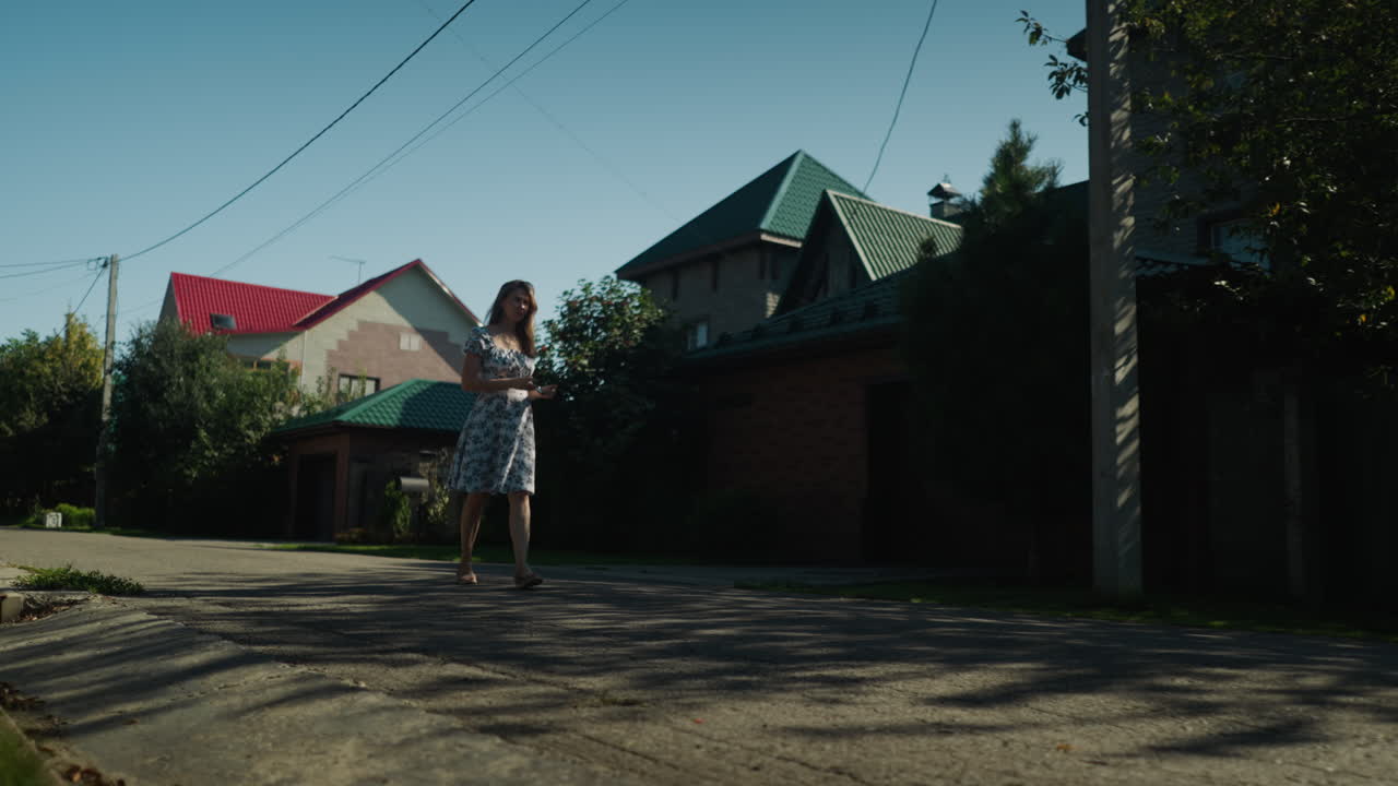 Young woman in floral dress pauses under tree shade to remove sunglasses and tuck them into dress while strolling leisurely along quiet sunlit suburban road lined with homes and greenery