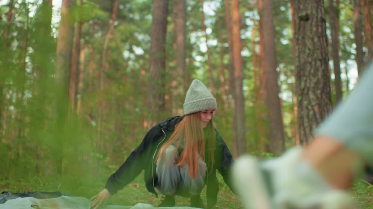 Young woman squatting on forest floor adjusting camping setup with focused posture as another person passes nearby, surrounded by pine trees in natural daylight