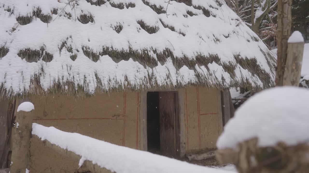 Rustic hut with a thatched roof stands covered in snow, nestled in a quiet winter village.