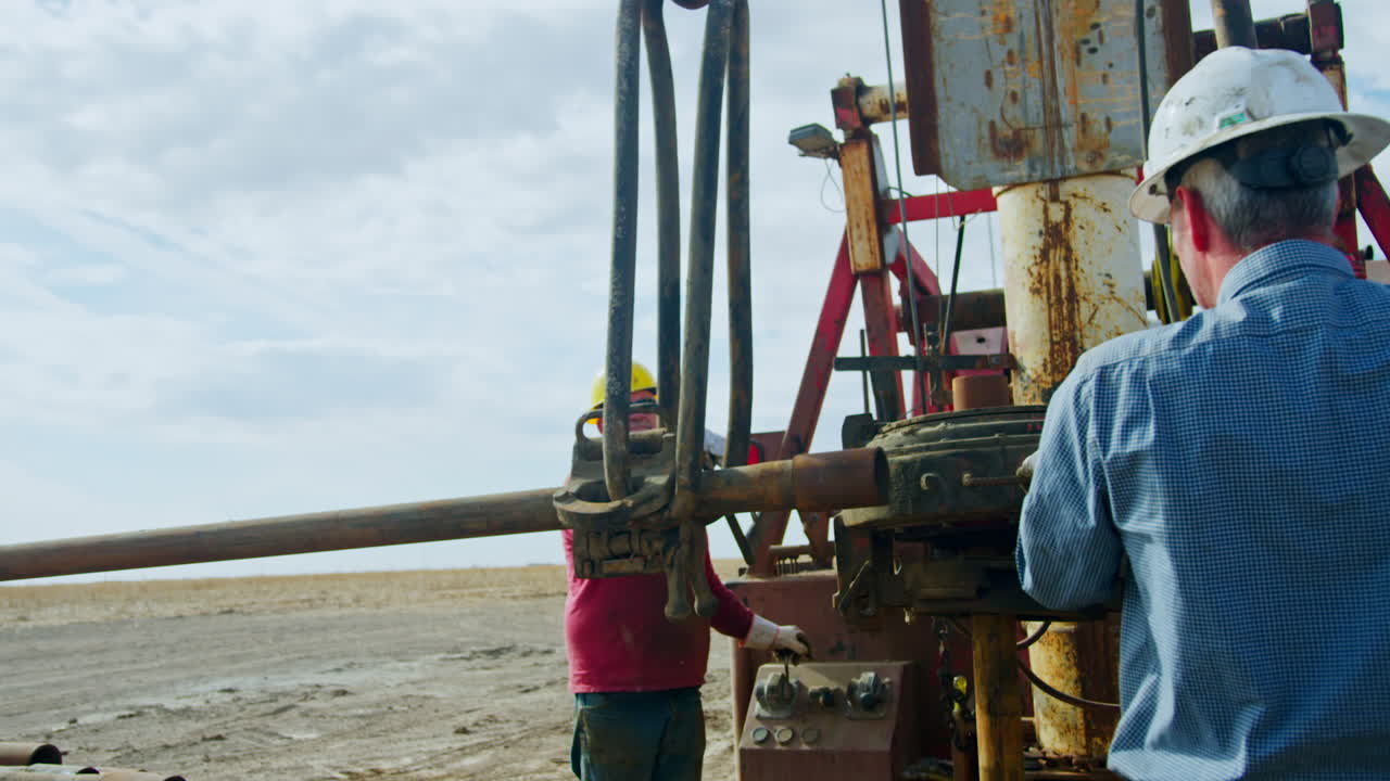 Two mature men installing equipment at industrial site for oil production. Metal pipe hanging on the hook is lifted up.