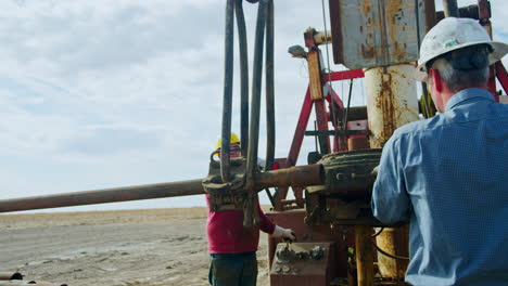 two mature men installing equipment at industrial site for oil production. metal pipe hanging on the hook is lifted up.