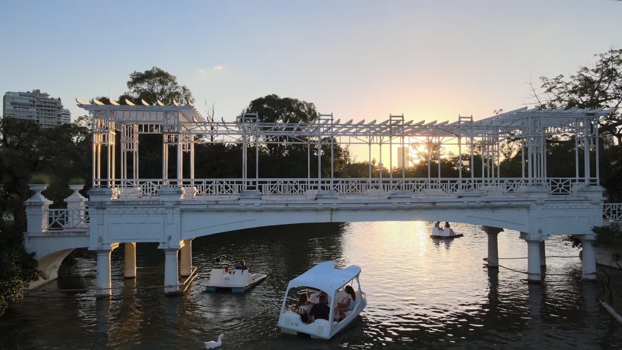 Aerial dolly in of people in boats sailing under white bridge in Rosedal gardens pond at sunset, Buenos Aires